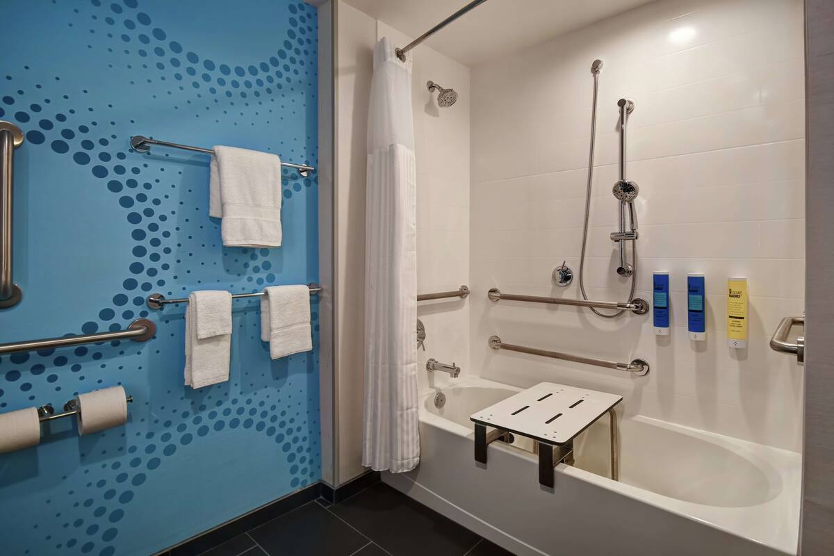 A bathroom with a blue wall, white fixtures, a bathtub with grab bars, a shower with a handheld showerhead, and a towel rack with towels.