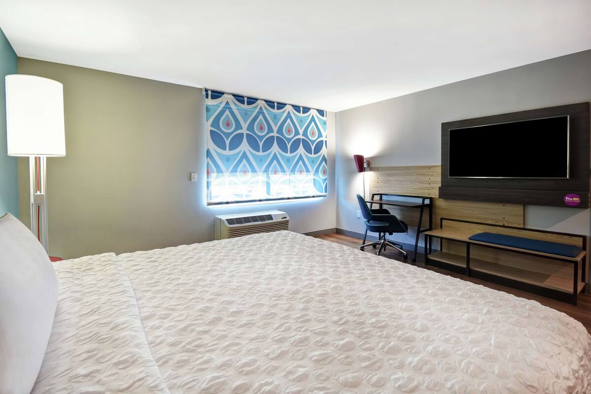A hotel room with a large bed, a desk with a chair, a flat-screen TV mounted on the wall, and a window with patterned curtains.