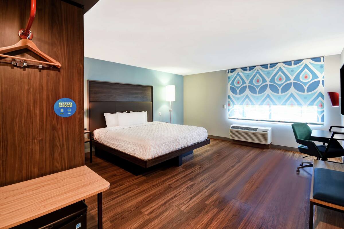 A hotel room with a bed, desk, chair, and a large window with a patterned curtain.