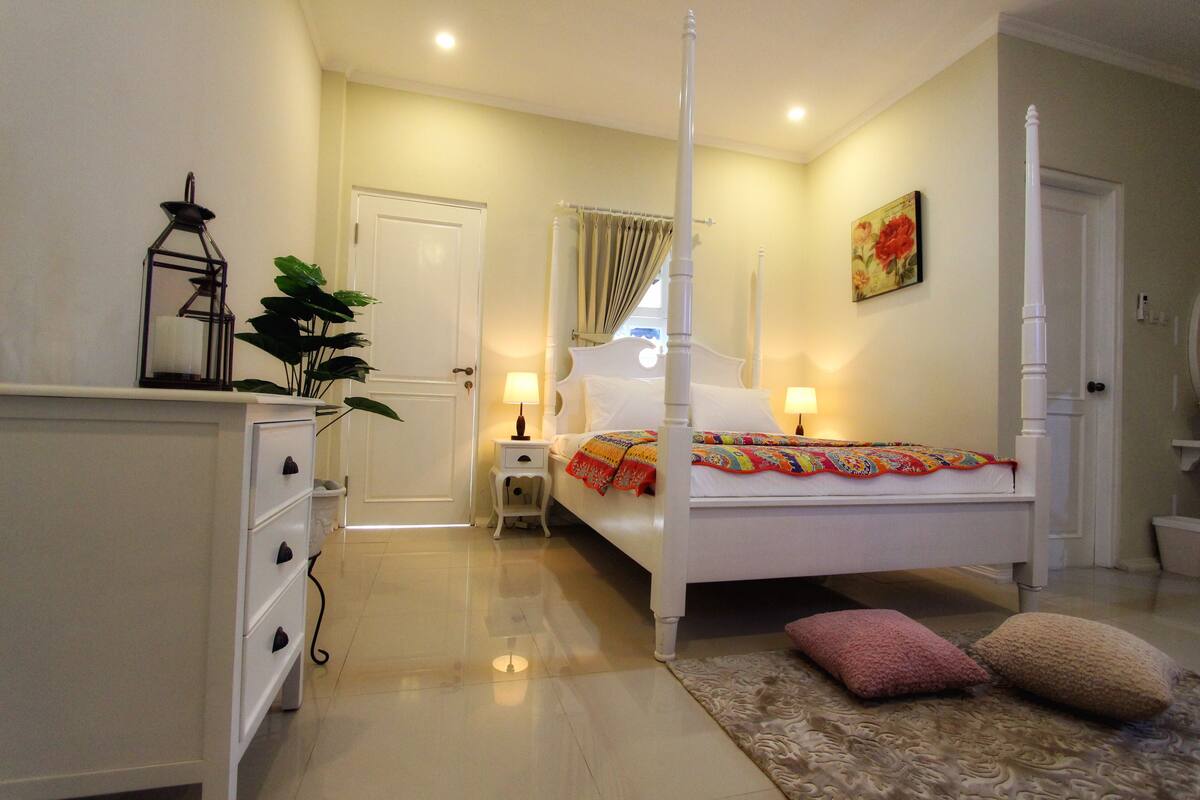 Une chambre à coucher avec un lit à baldaquin, des tables de chevet avec des lampes, une table de nuit, une commode, une plante en pot et un sol recouvert d’une moquette.