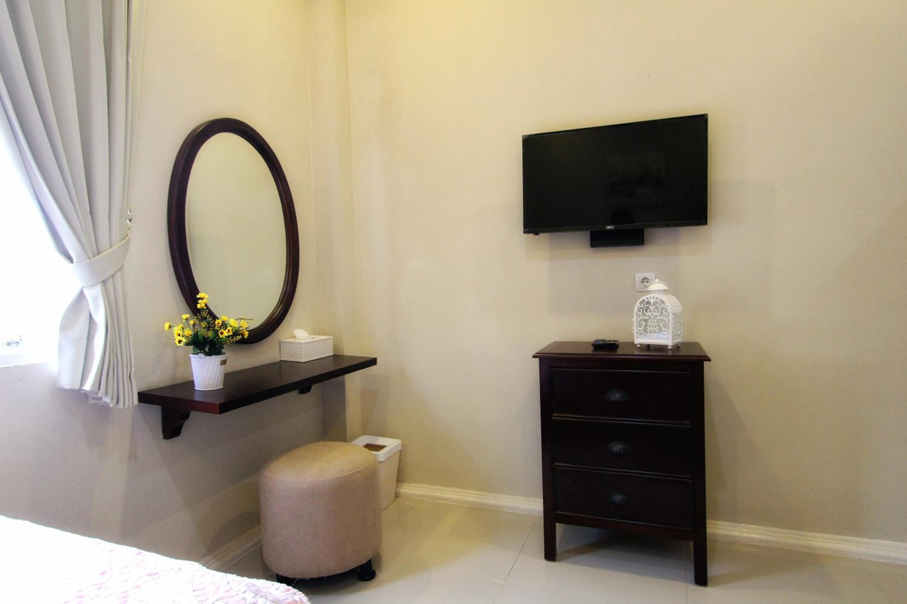 Un dormitorio con cama, mesita de noche con espejo, cómoda, televisor montado en la pared y ventana con cortinas.
