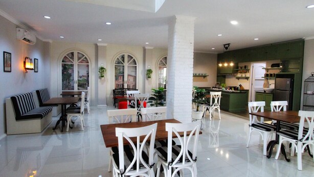 A modern dining area with white chairs and wooden tables, a kitchen with green cabinets, and a seating area with a bench and chairs. - Omah Madam Bed & Breakfast (Semarang)