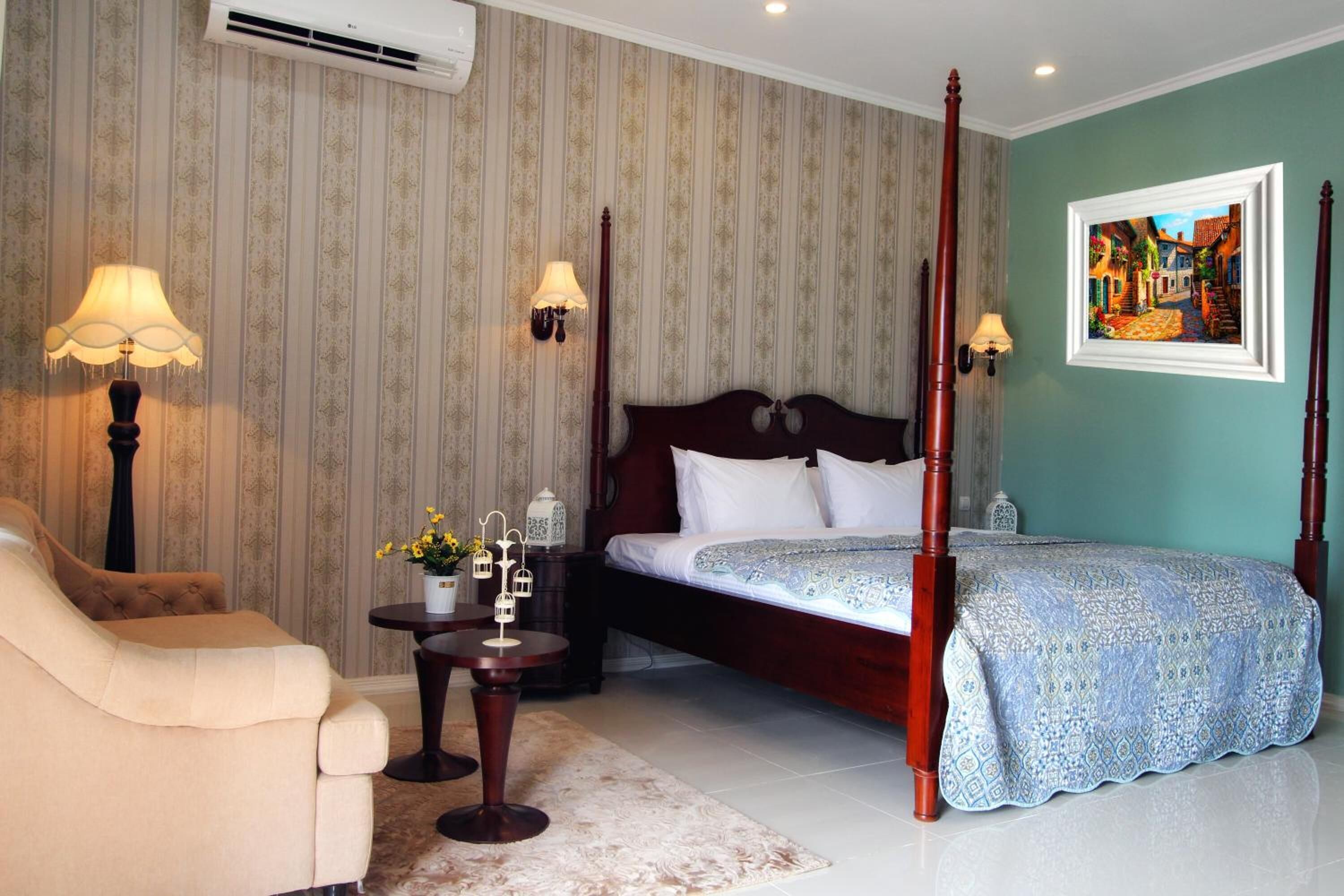 Een hemelbed met een donker hoofdeinde, een beige fauteuil, een klein rond tafeltje met een lamp en een schilderij aan de muur.