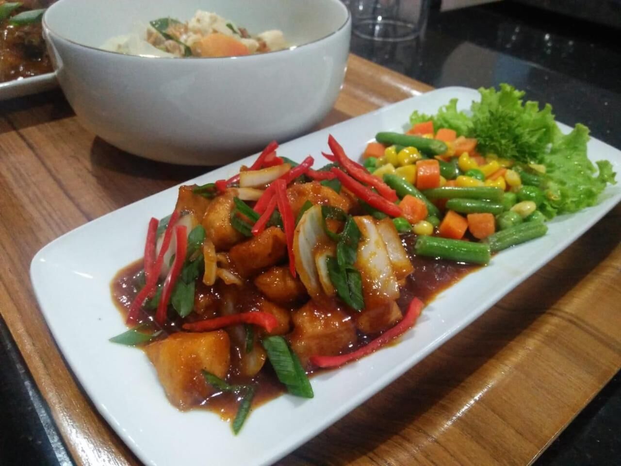Een rechthoekig bord met een portie roerbakgroenten en tofu, versierd met rode en groene paprika’s, geserveerd op een houten tafel.