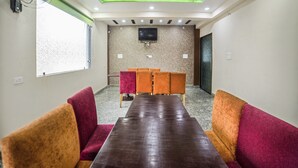 A conference room with a long table, orange and red upholstered chairs, a ceiling fan, recessed lighting, a TV mounted on the wall, and a window with blinds.