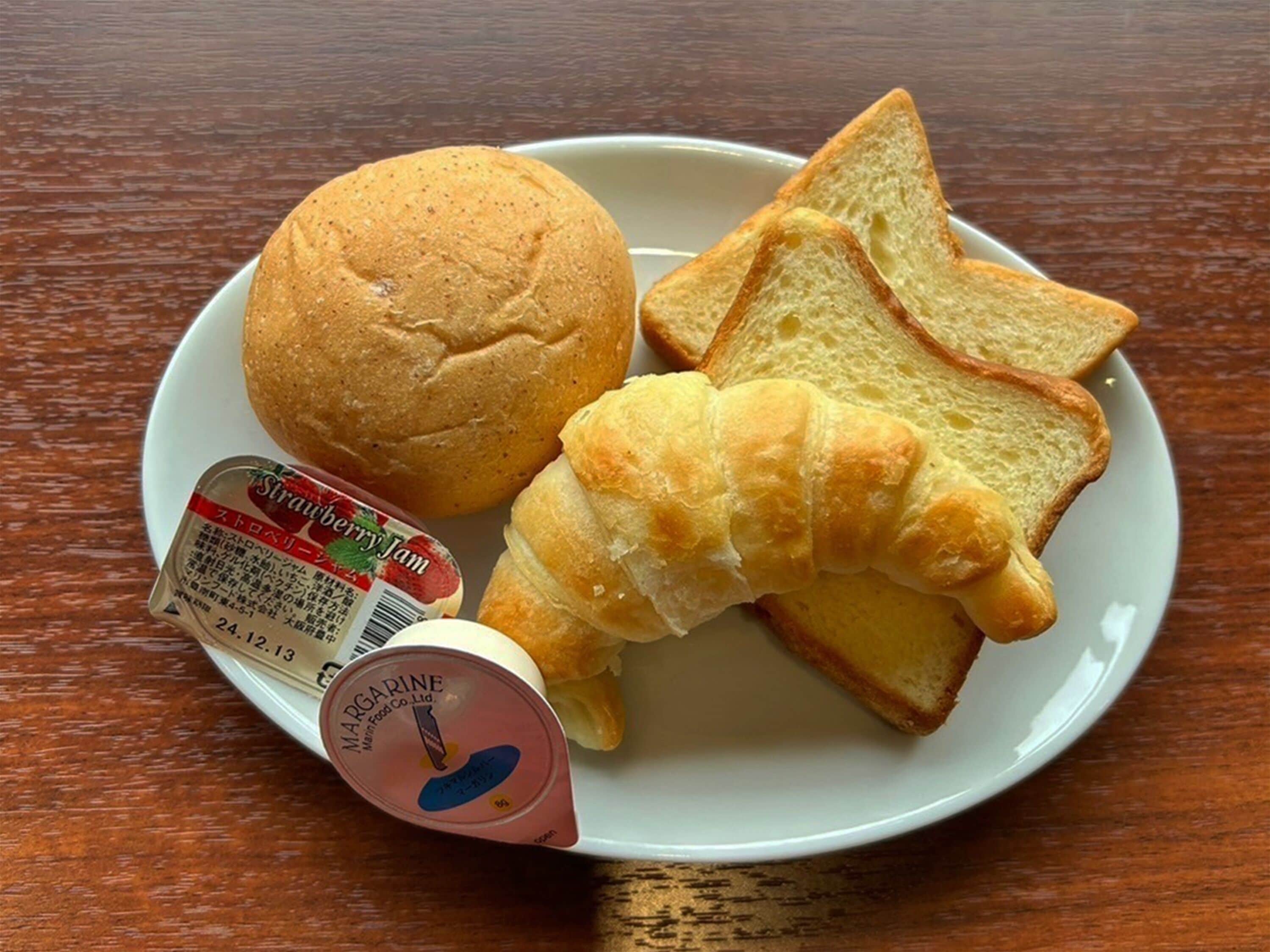 A plate with a bread roll, croissant, and slices of bread, along with a jar of strawberry jam and a margarine container.