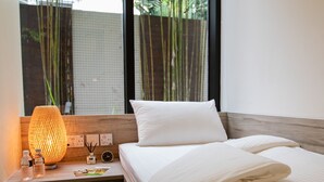 A neatly made bed with white linens, a wooden nightstand with a textured lamp, and a window with bamboo outside. - JetQuay Suites (Singapore)