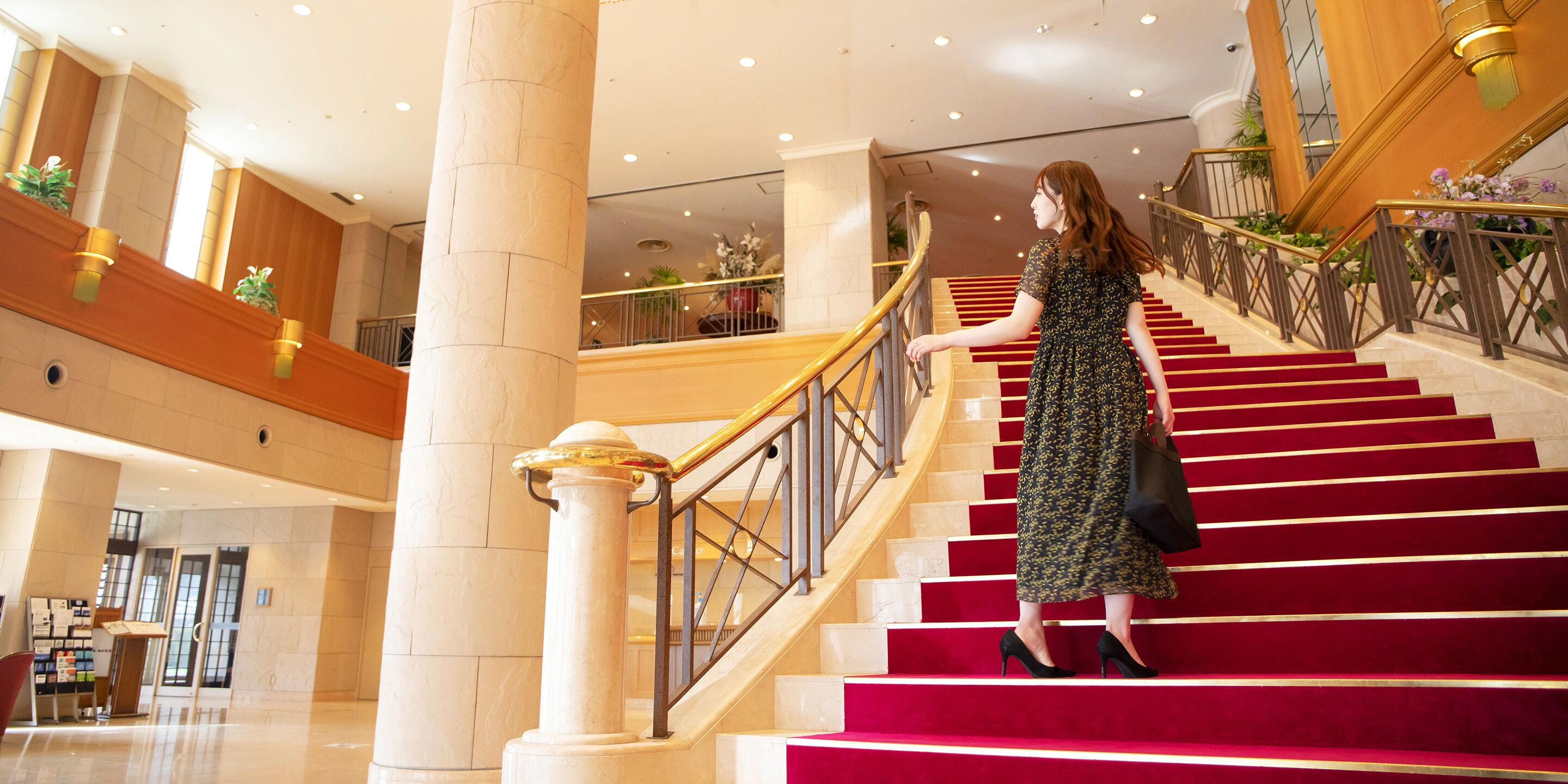 A woman descending a red carpeted staircase in a hotel lobby with marble columns and a wooden railing.