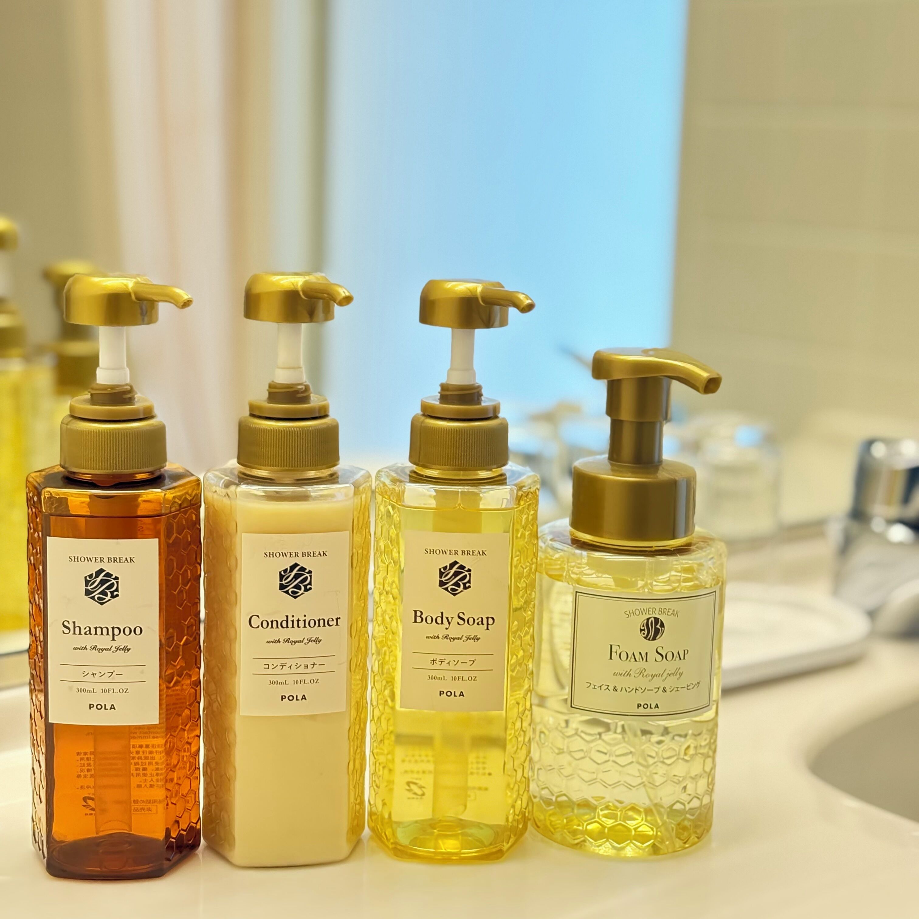 Neljä suihkugeelin ja muiden suihkutuotteiden pulloa annostelupumpuilla, sisältäen shampoota, hoitoainetta, nestesaippuaa ja vaahtoavaa saippuaa, on asetettu riviin kylpyhuoneen tasolle.