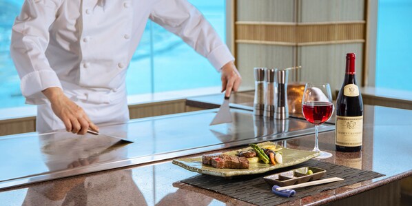 A chef preparing food at a counter with a bottle of wine and a glass of red wine.