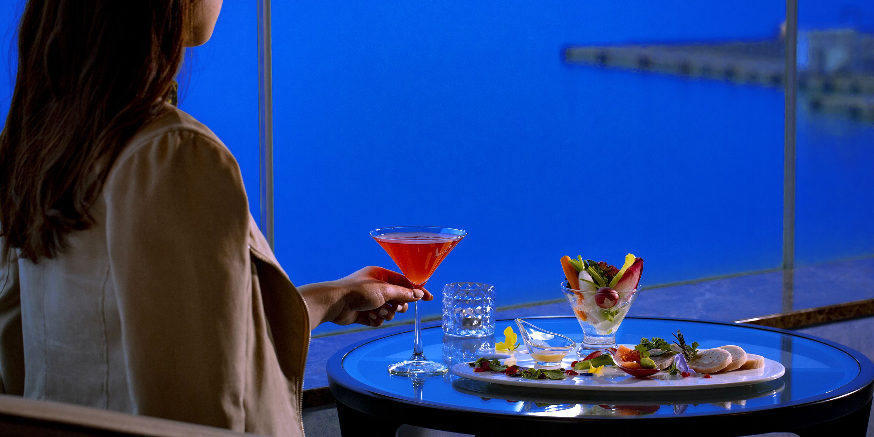 A person holding a martini glass at a table with a salad and a plate of food.