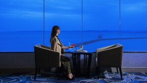Een vrouw zit aan een tafel met uitzicht op zee en een zeilboot.