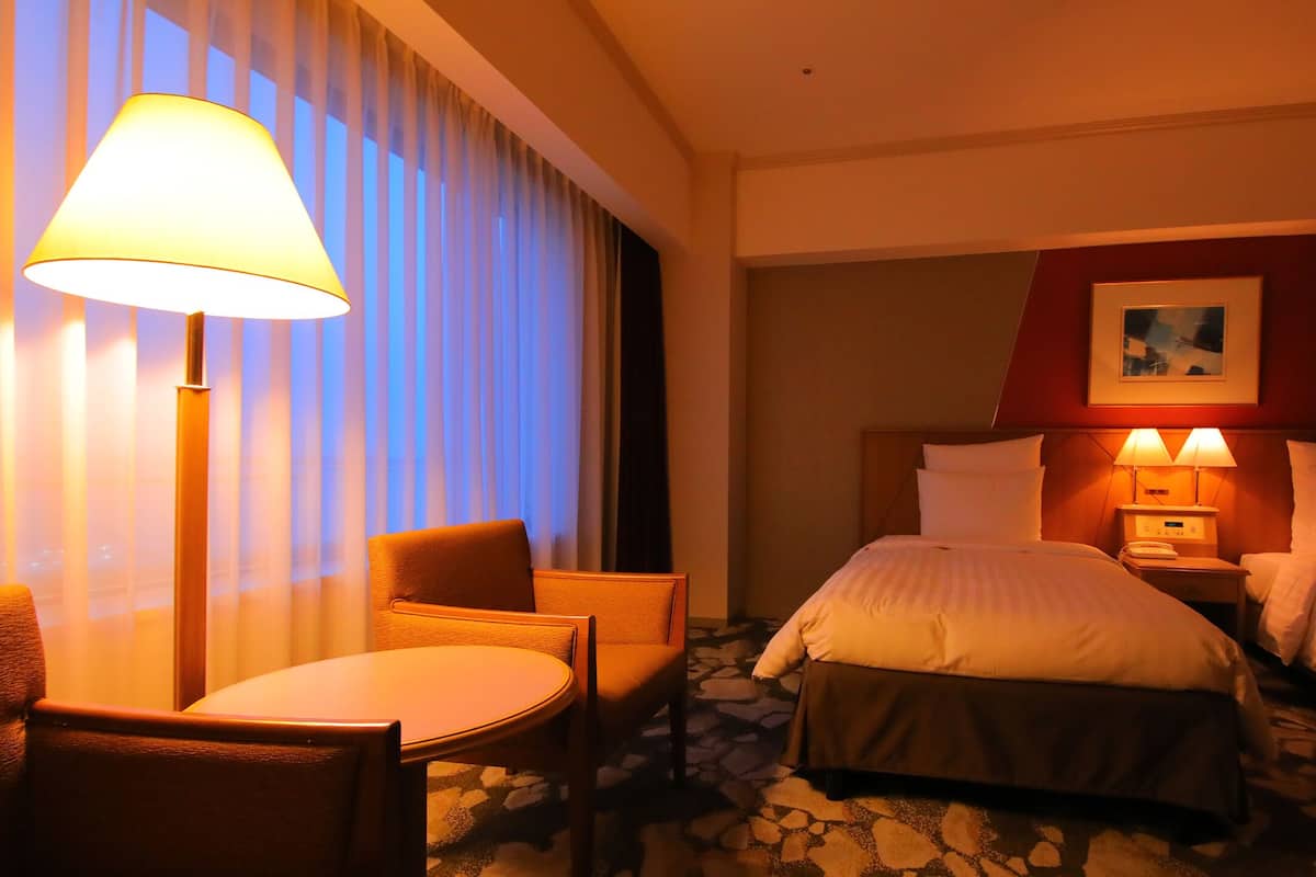 Ein Hotelzimmer mit einem Bett, zwei Nachttischen, einem Sessel, einem Tisch und einem fenster mit Vorhängen.
