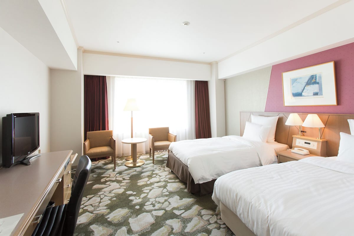 Ein Hotelzimmer mit zwei Betten, einem Schreibtisch, einem Fernseher, einem kleinen Tisch und einem Sessel.