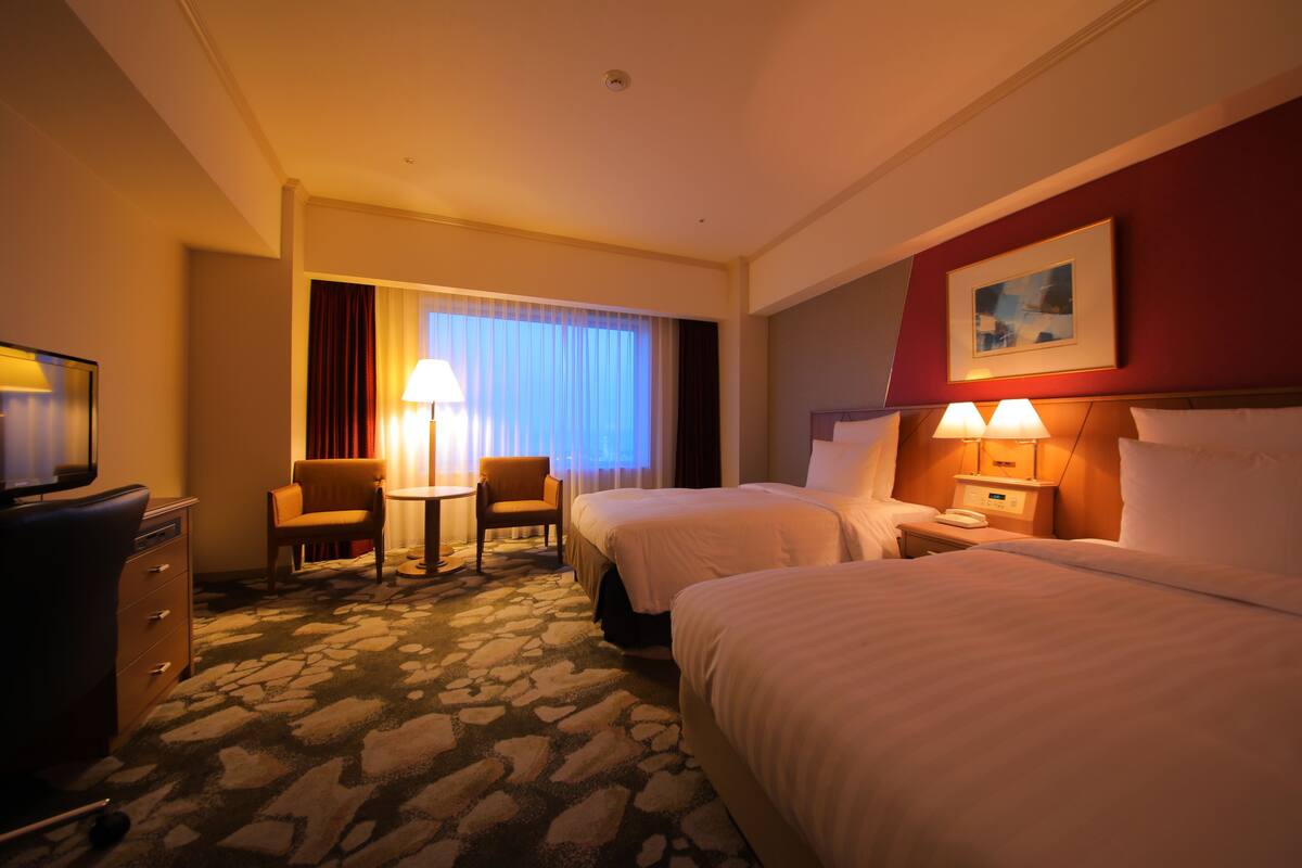 Ein Hotelzimmer mit zwei Betten, einem Schreibtisch, einem Stuhl, einer Lampe und einem Fernseher.