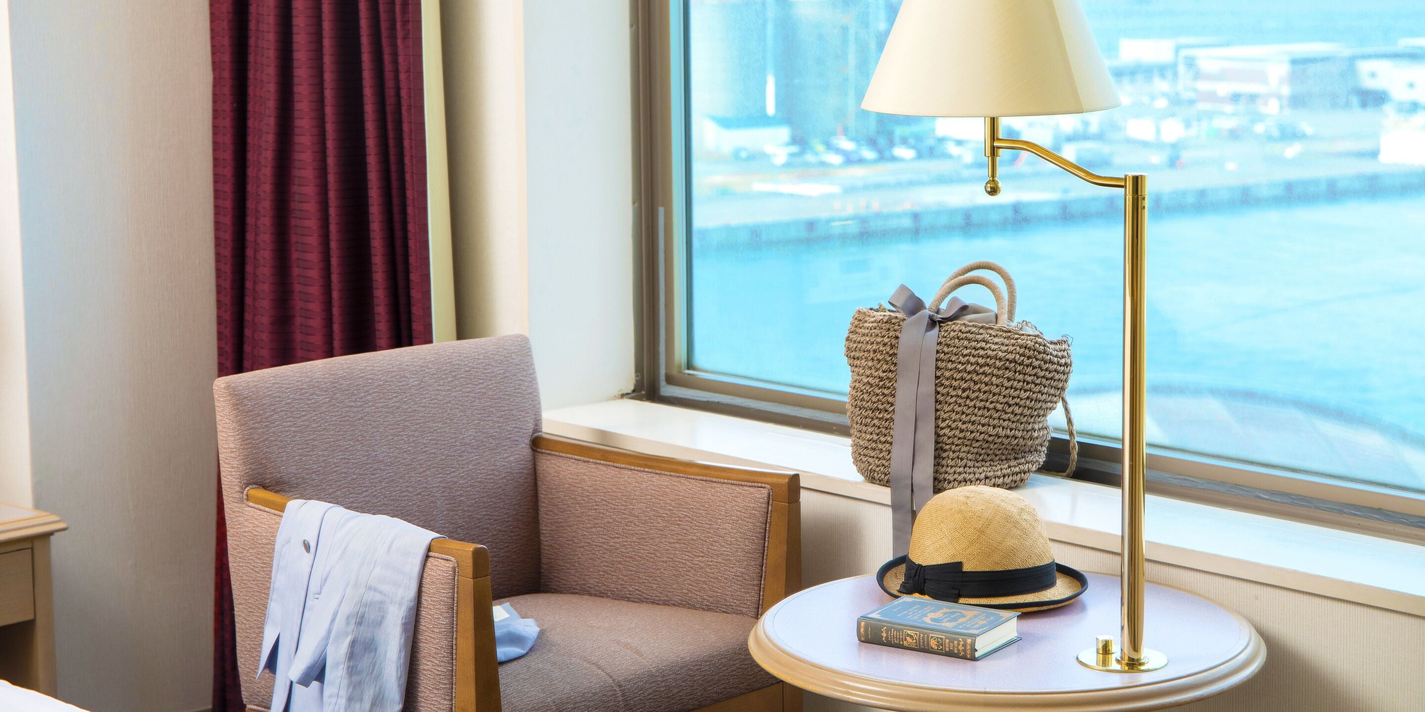 A hotel room with a view of a body of water, a chair, a lamp, a hat, and a book on a small table.