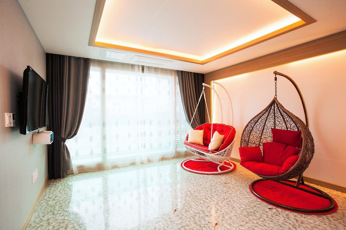 Ein modernes Hotelzimmer mit großem Fenster, einem Flachbildfernseher, einem roten Hängekorbsessel und einem roten Sofa.