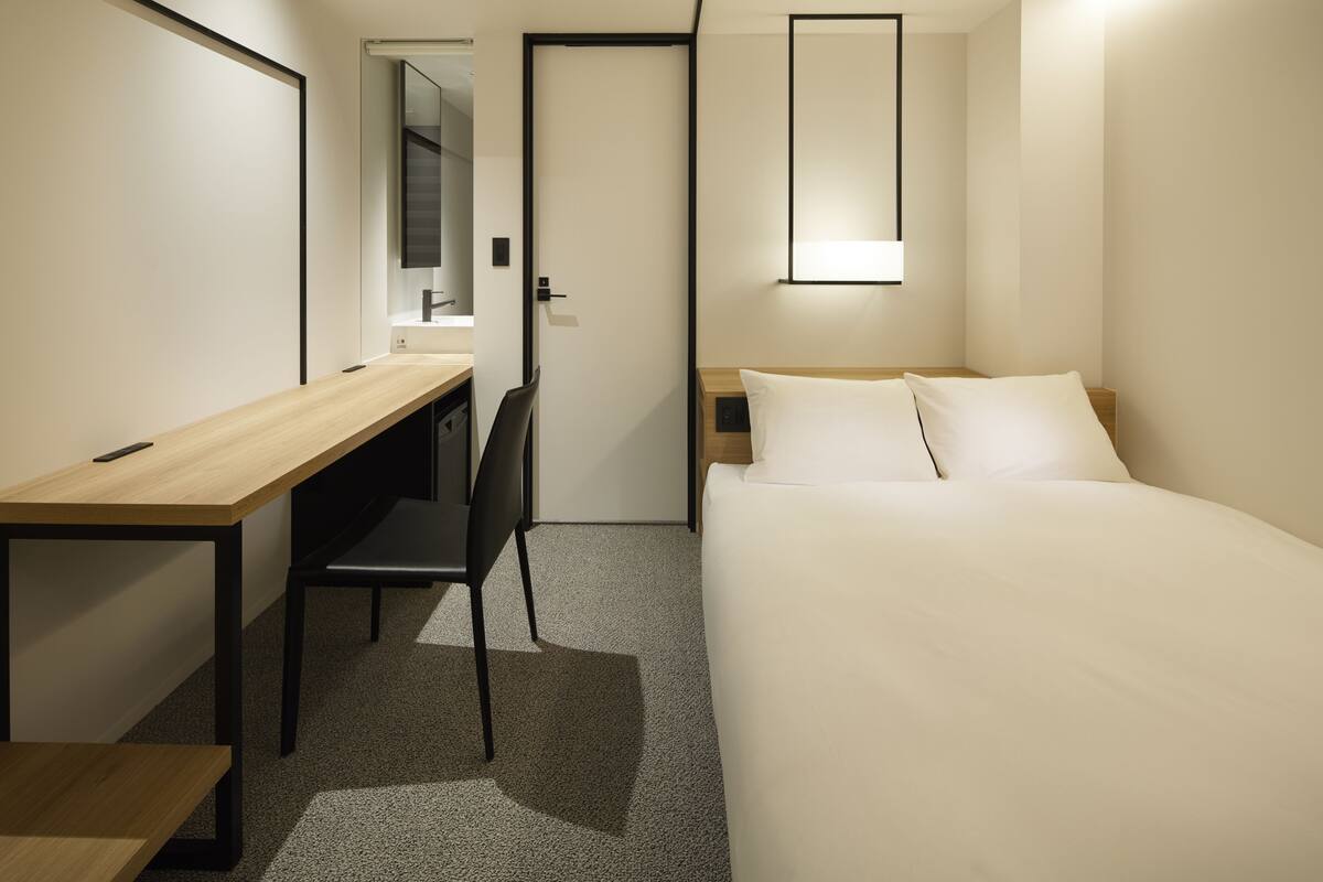 Een moderne hotelkamer met een houten bureau, een zwarte stoel, een bed met witte lakens en een nachtkastje met een lamp.
