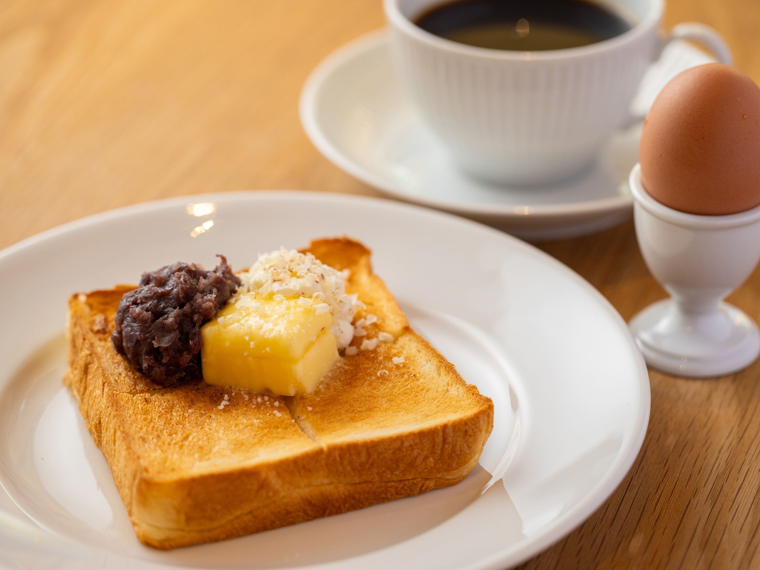 A breakfast plate with toast, butter, and jam, a cup of coffee, and a boiled egg on a table.