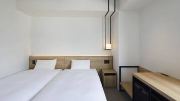 A modern hotel room with two single beds, a wooden headboard, a bedside table, a desk, and a wall-mounted light fixture.