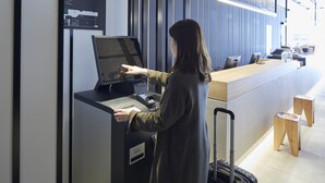Eine Frau benutzt eine Selbstbedienungskiosk in einer modernen Hotelhalle mit einem Holzerrepsionsschalter und freier Sicht auf den Empfangsbereich.