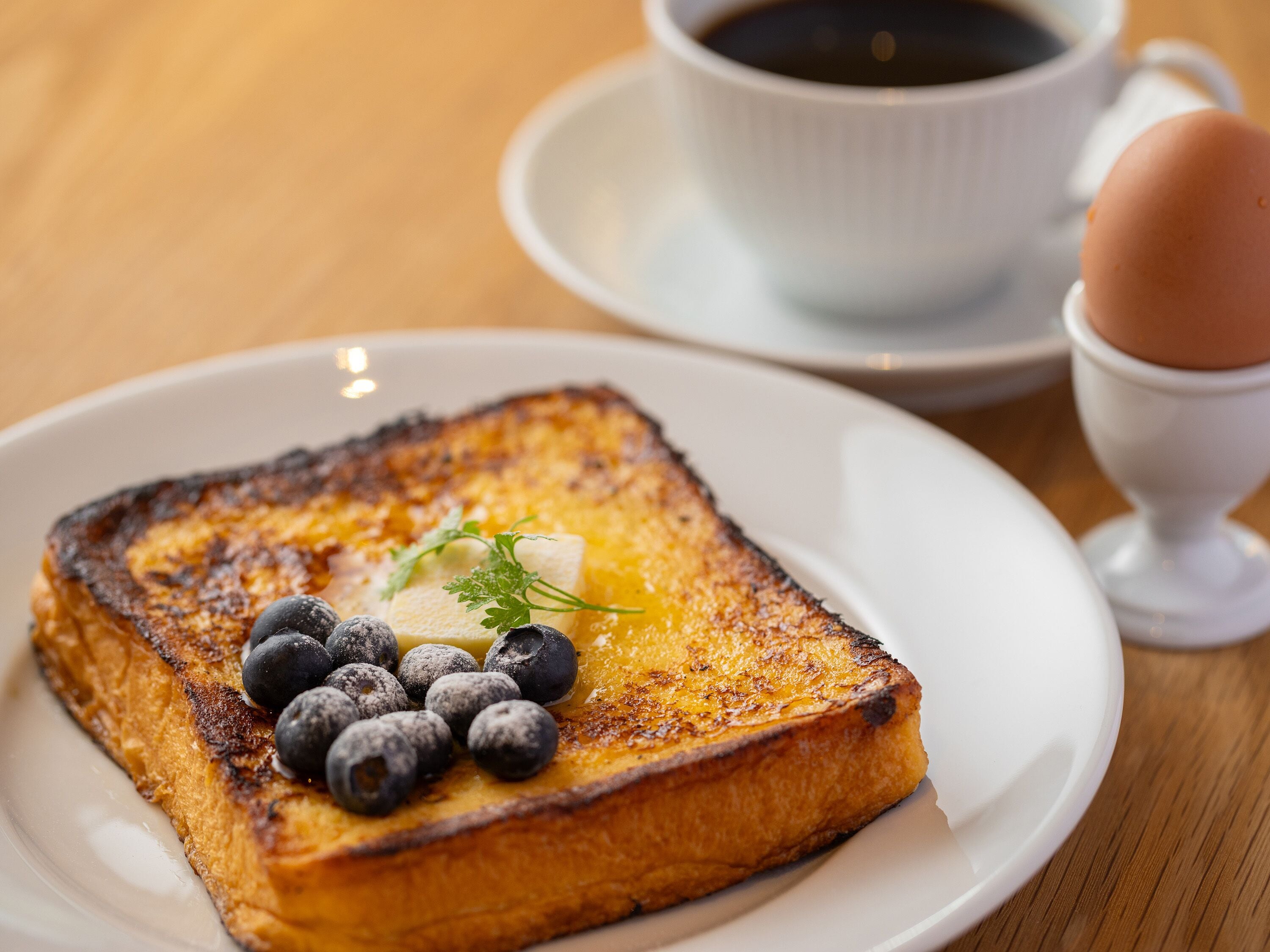 Une assiette de pain perdu aux myrtilles, garnie d’une petite noix de beurre, accompagnée d’une tasse de café et d’un œuf dans une soucoupe.