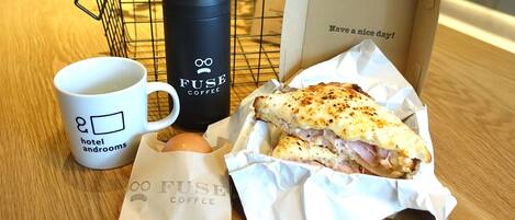 Una mesa con una taza de café, una botella de agua negra, un sándwich, una bolsa pequeña y una caja de cartón con un mensaje.