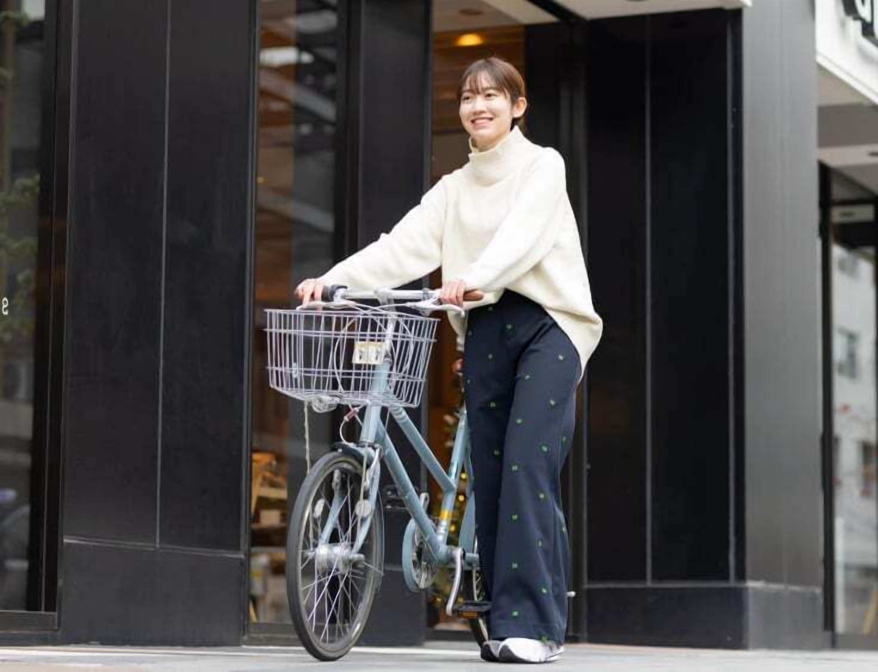 Una persona amb bicicleta i cistella, de peu davant d'un edifici amb grans finestres.