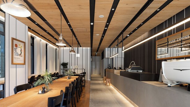 Ein modernes Interieur mit einem langen Holztisch, schwarzen Stühlen, Pendelleuchten und einer Kaffeebar mit dem Schild "FUSE COFFEE".