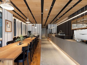 Un intérieur moderne avec une longue table en bois, des chaises noires, des luminaires suspendus et un coin café agrémenté d’une enseigne indiquant « FUSE COFFEE ».