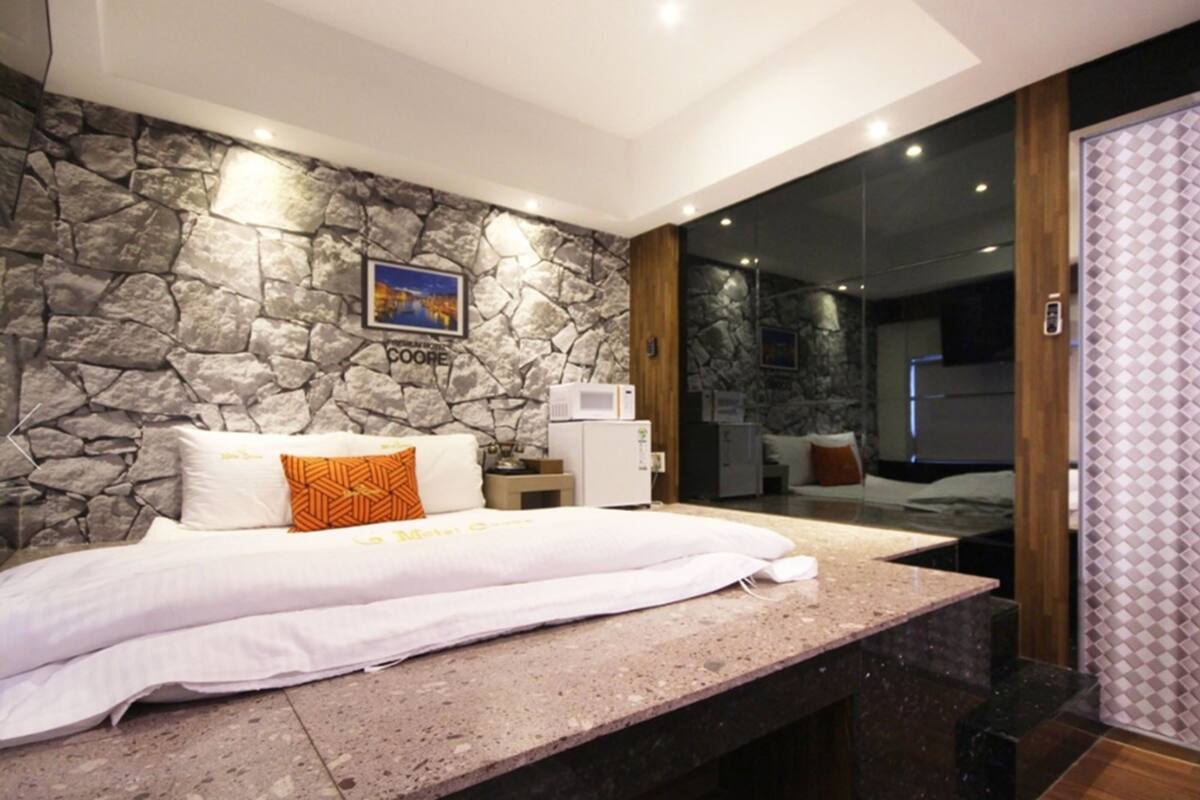 A modern bedroom with a stone wall, a bed with white linens, a small bedside table, and a large mirror reflecting the room.