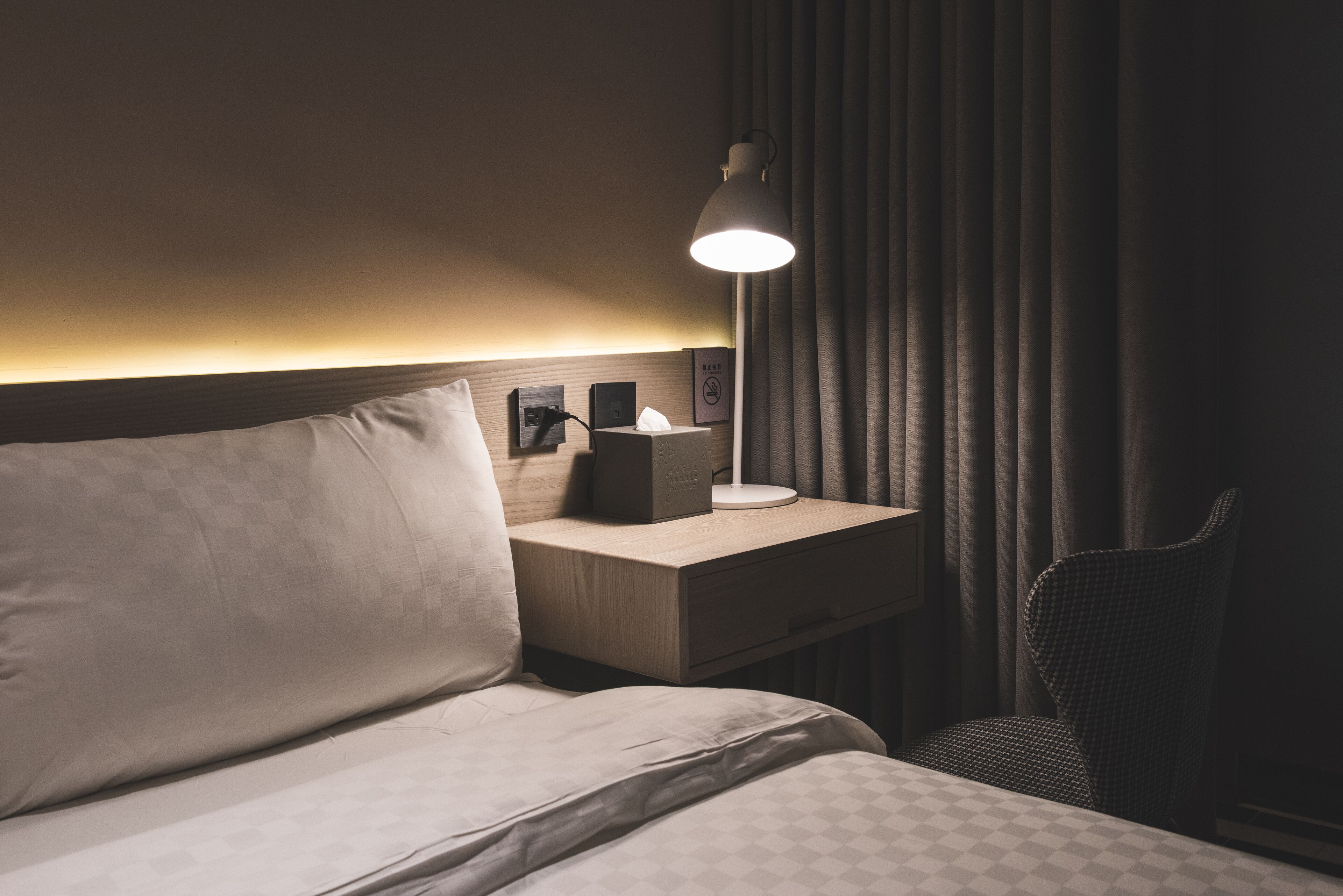 A hotel room with a bed, a bedside table with a lamp, a tissue box, and a chair.