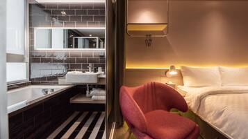 Een moderne hotelkamer met een groot bed, een rode fauteuil, een badkamer met een grote spiegel en een zwart-wit gestreept vloerkleed.