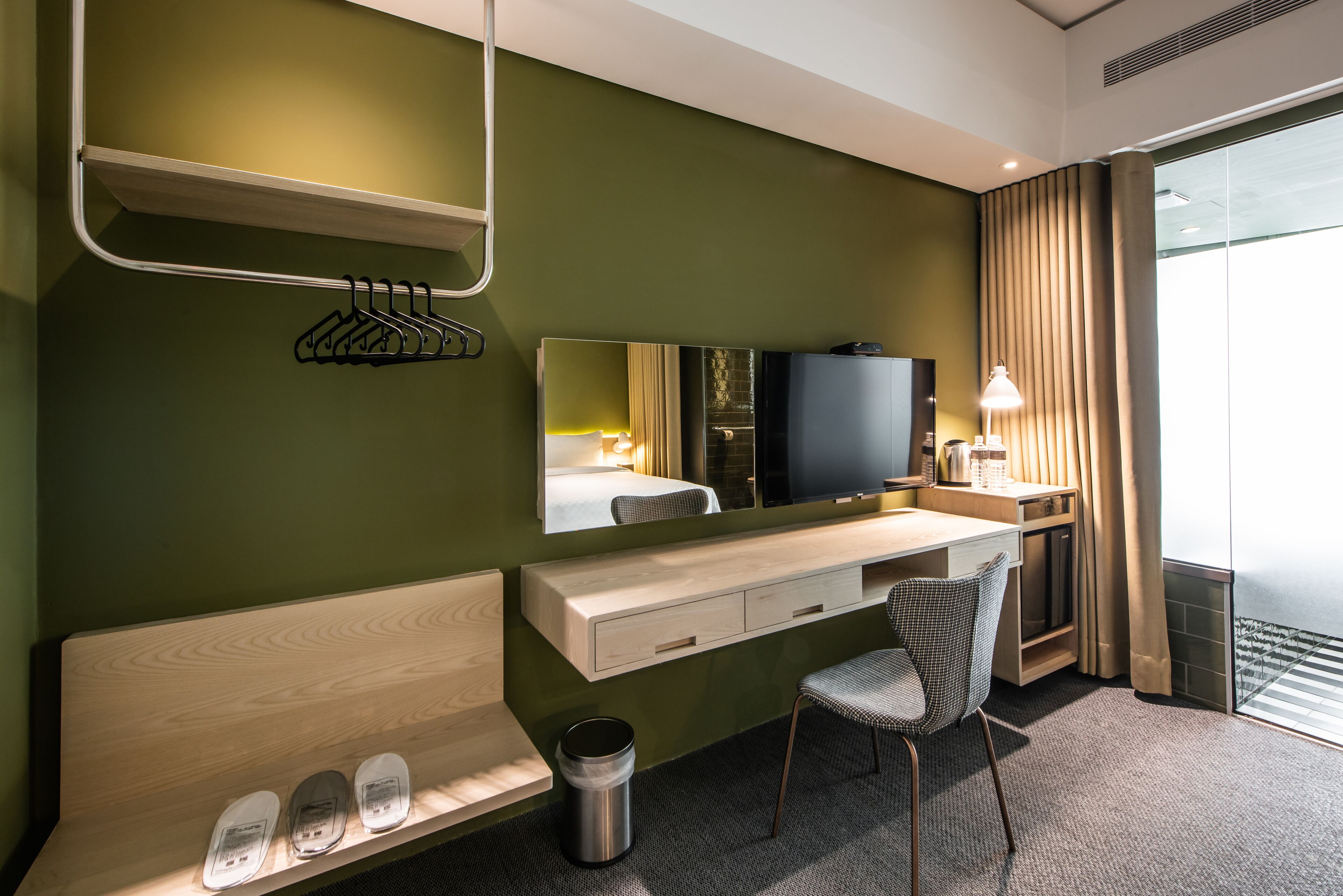 Habitación de hotel con un escritorio de madera, una silla, un televisor, un espejo y una ventana con cortinas.