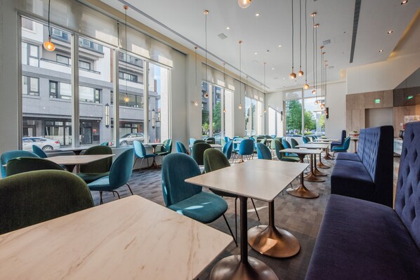 A modern restaurant interior with large windows, blue and green chairs, and a mix of table styles. - Hotel Ramble (Taichung)