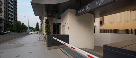 Entrada moderna de un edificio con un letrero que dice 'Welcome', señal de estacionamiento que indica una capacidad de 1.75 metros, y un camino de acceso con puerta de reja.