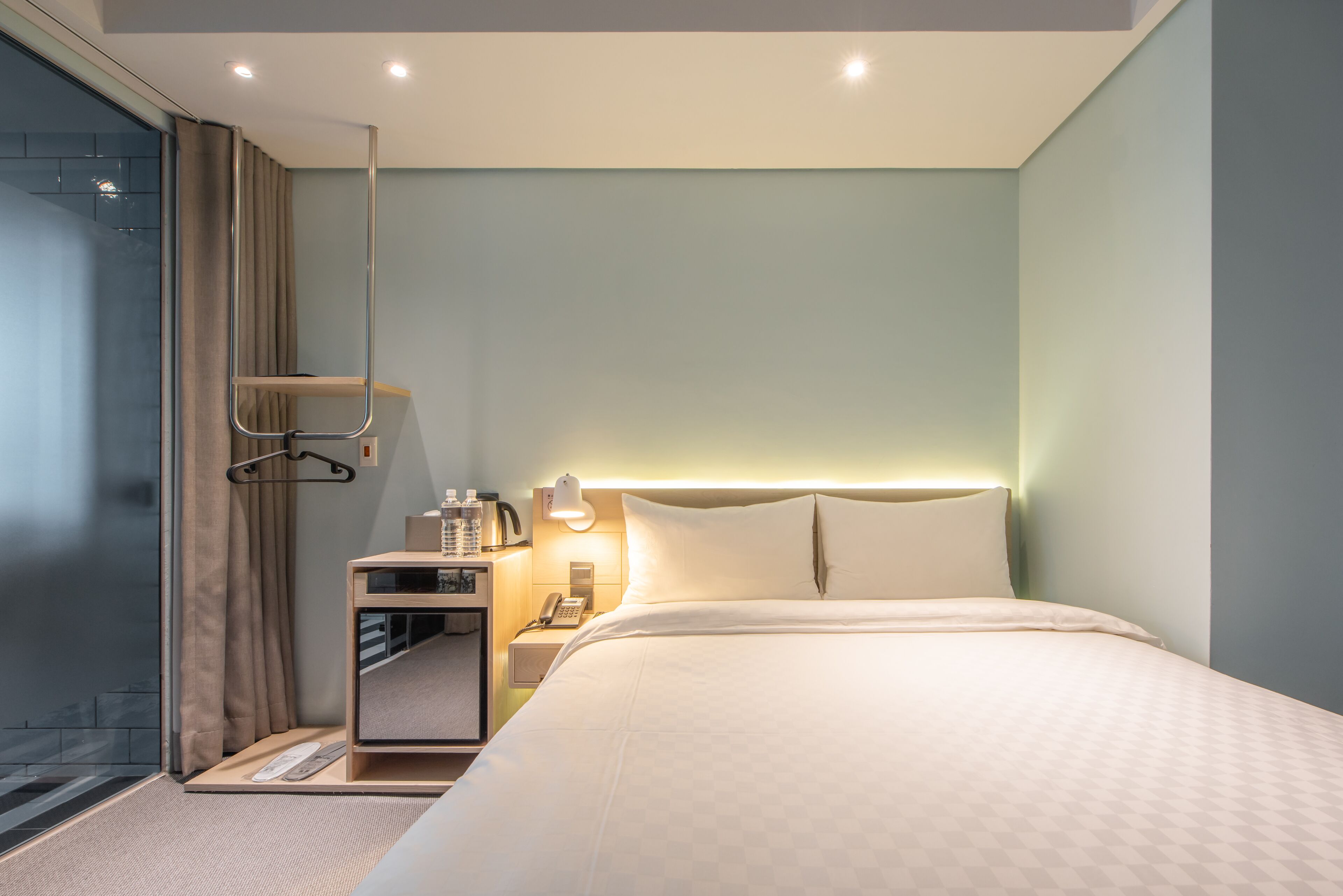 Una habitación de hotel moderna con una cama grande, una mesita de noche con lámpara, un teléfono y un estante pequeño con artículos de aseo.