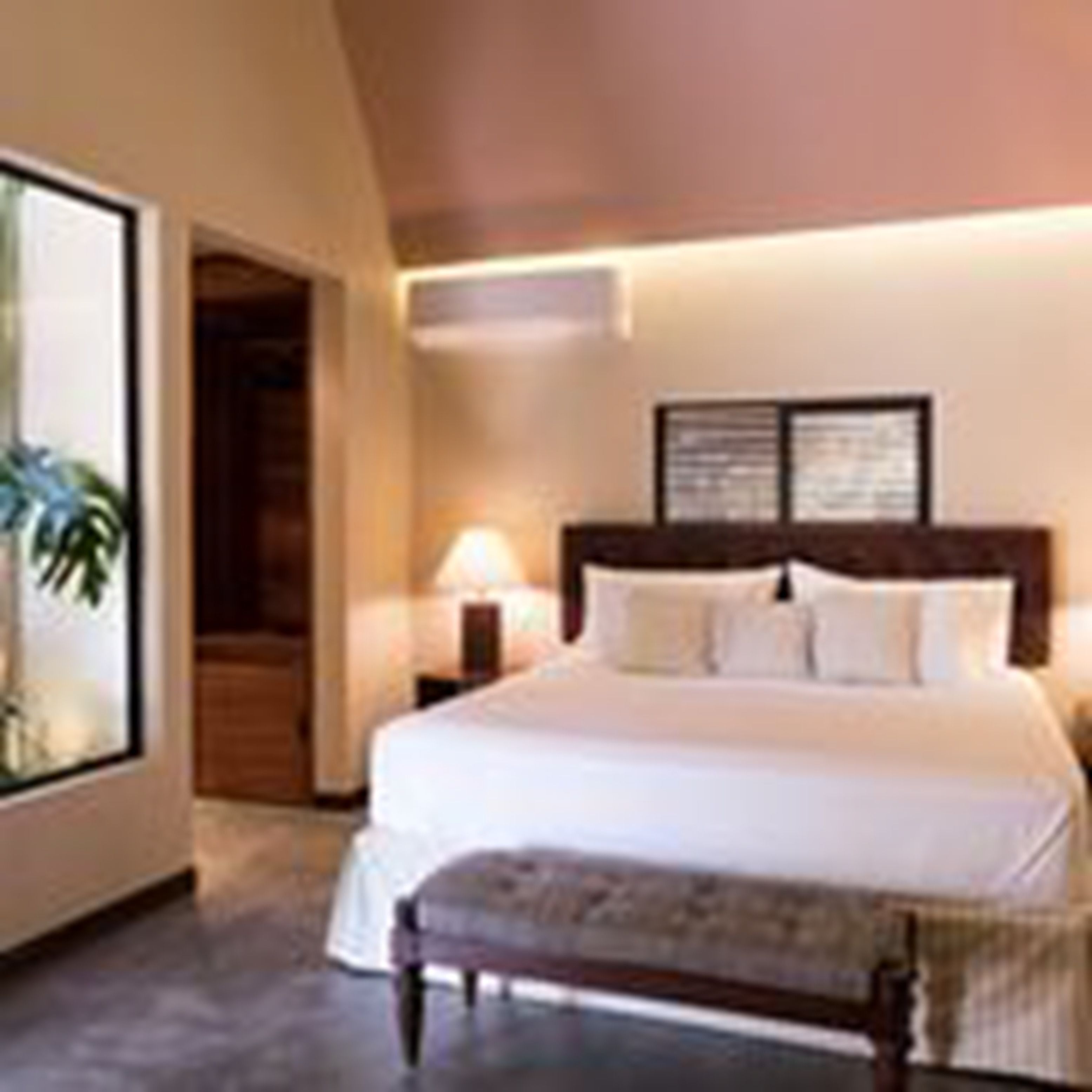 A bedroom with a large bed, a nightstand, a lamp, and a window with a view of greenery.