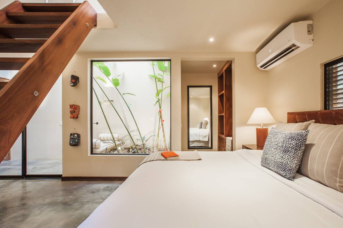 Ein Schlafzimmer mit einem großen Bett, einer Holztreppe, einem Fenster mit Pflanzen, einer Nachttischlampe und einer Klimaanlage.