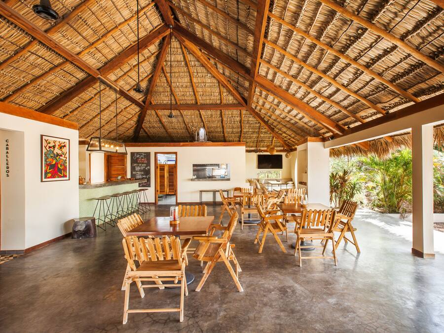 A spacious indoor dining area with wooden tables and chairs, a thatched roof ceiling, and a view of greenery outside.