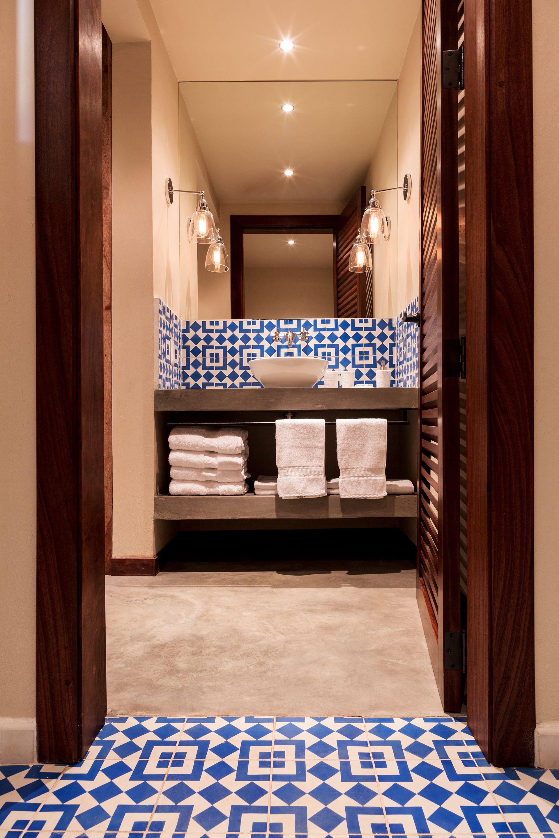 Un pasillo estrecho que conduce a un baño con suelo de baldosas blancas y azules, un tocador de madera con lavamanos y dos lámparas fijadas a la pared.
