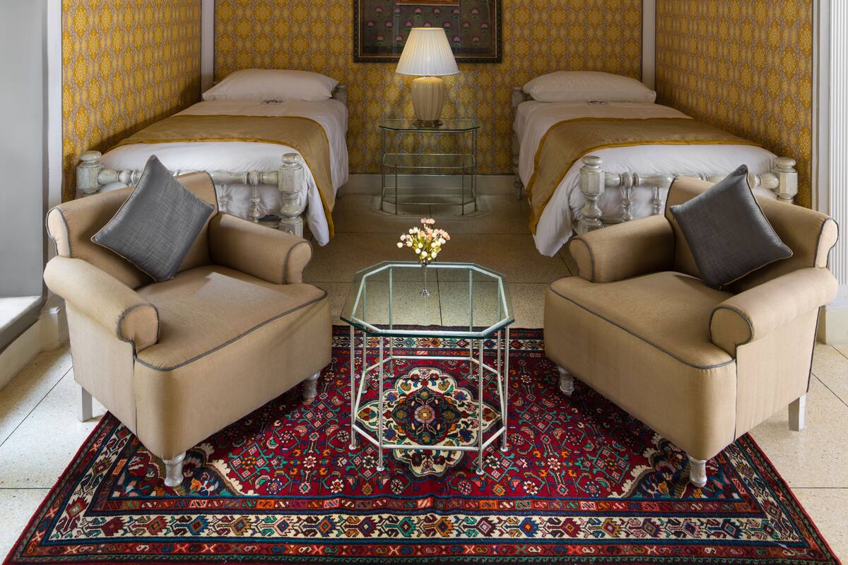 Habitación de hotel con dos camas, un candelabro, una mesa con un florero y papel tapiz estampado.