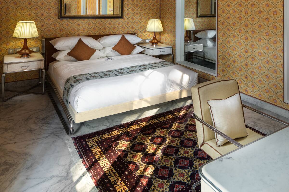 Habitación de hotel con una cama grande, dos mesitas de noche con lámparas, una araña, un espejo y una alfombra estampada.