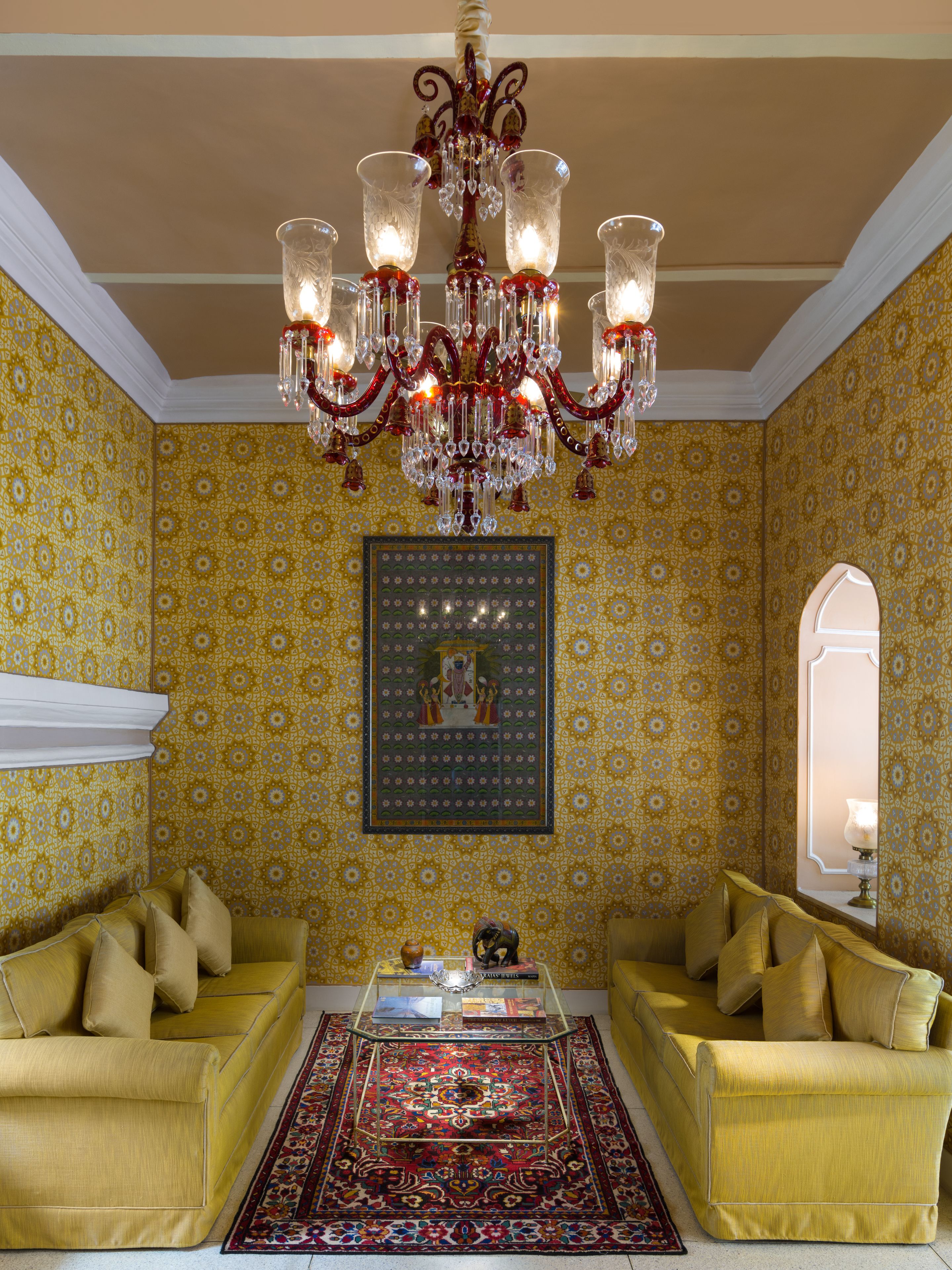 A room with yellow patterned wallpaper, a chandelier, two yellow sofas, a coffee table, and a framed picture on the wall.
