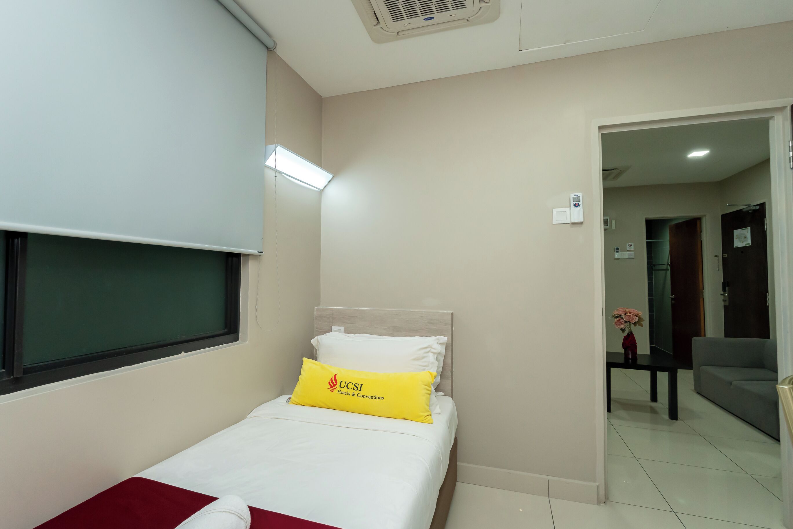 A hotel room with a bed, a window, a bedside table with a vase of flowers, and a door leading to another room.