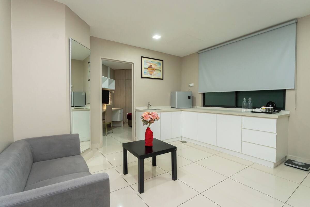 A modern living room with a grey sofa, a small black table with a vase of flowers, a kitchen area with white cabinets, a microwave, and a scale on the floor.