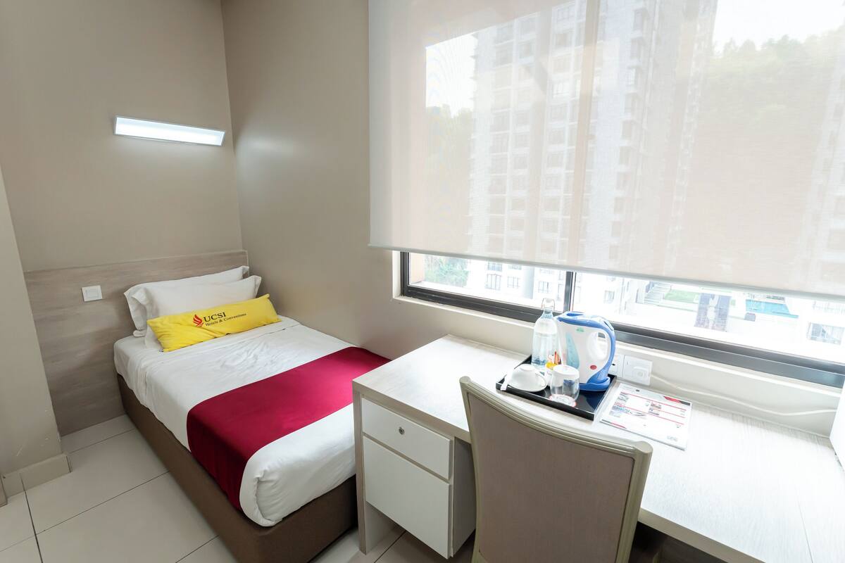 A hotel room with a bed, a desk, a chair, a window, and a view of buildings outside.