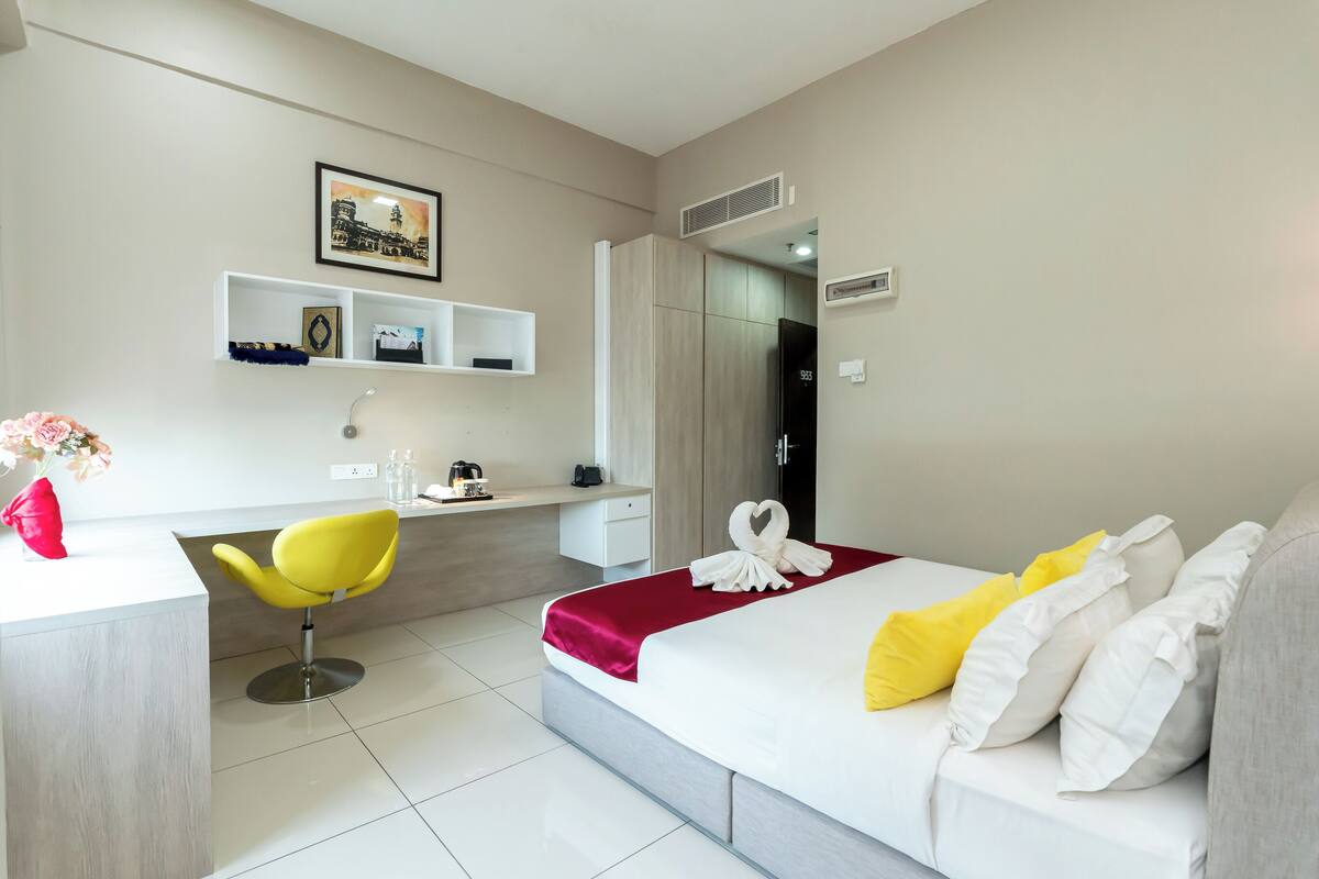 A hotel room with a bed, a desk, a chair, a small shelf, and a picture on the wall.