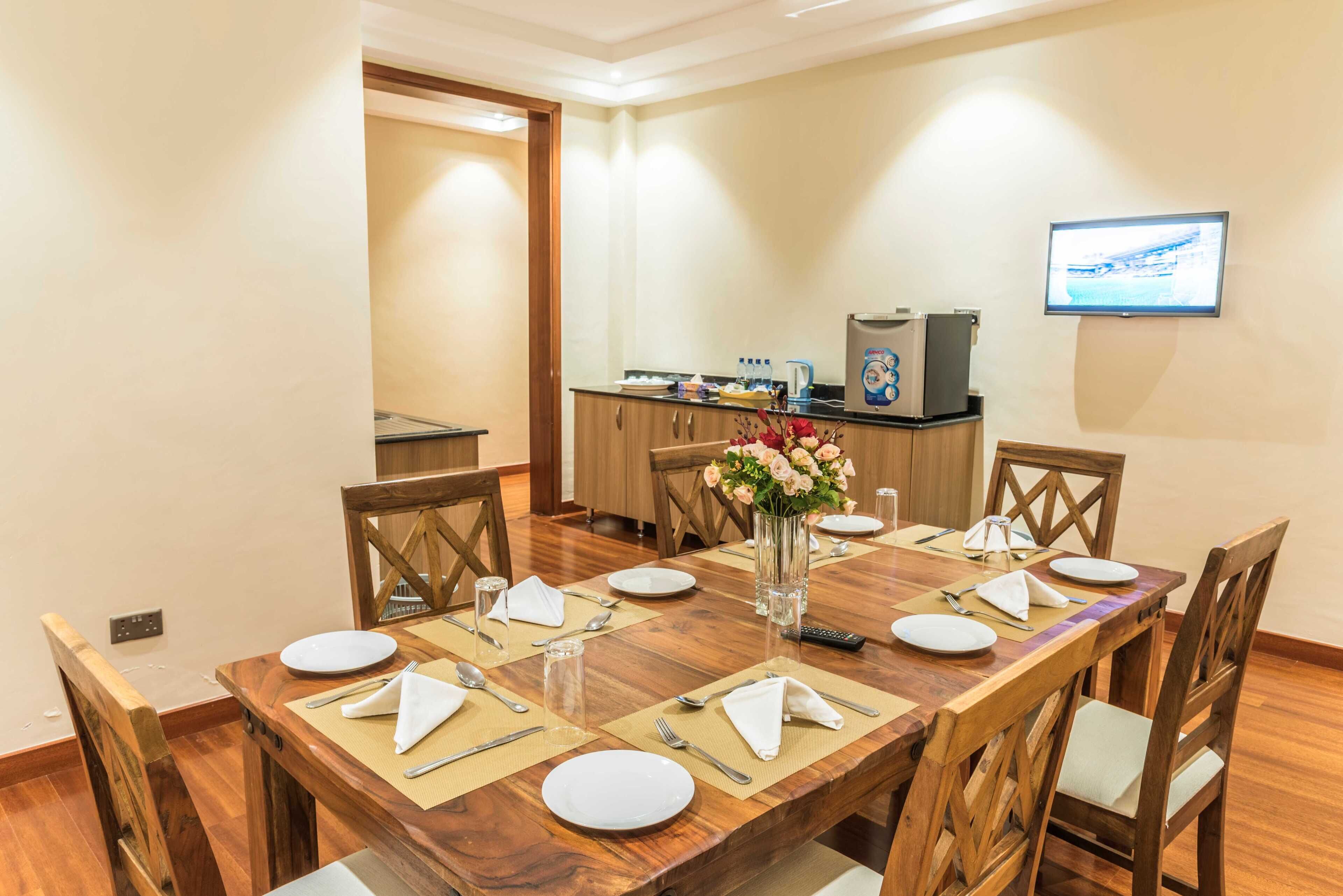 Un comedor con una mesa de madera dispuesta para seis personas, con platos, cubiertos y servilletas. Un jarrón con flores está sobre la mesa, y un televisor está montado en la pared.