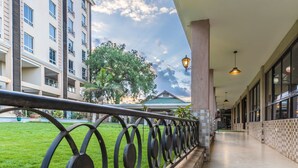 A corridor with a metal railing, a green lawn, and a building with multiple windows and balconies. - Three Steers Hotel (Meru)